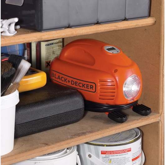BLACK+DECKER ASI200 oranje draagbare luchtcompressor met spiraalkabel, opgeborgen op een houten plank tussen gereedschap en verfblikken.