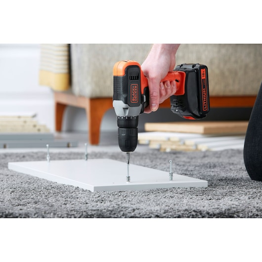 Perceuse sans fil BLACK+DECKER BCD001C2 utilisée pour assembler un panneau de meuble blanc sur un tapis gris, gros plan.