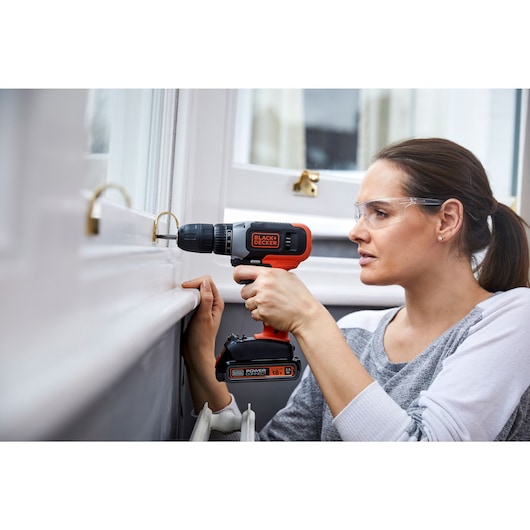 Personne utilisant la perceuse sans fil BLACKDECKER BCD001C2 sur un cadre de fenêtre, marque et batterie visibles.