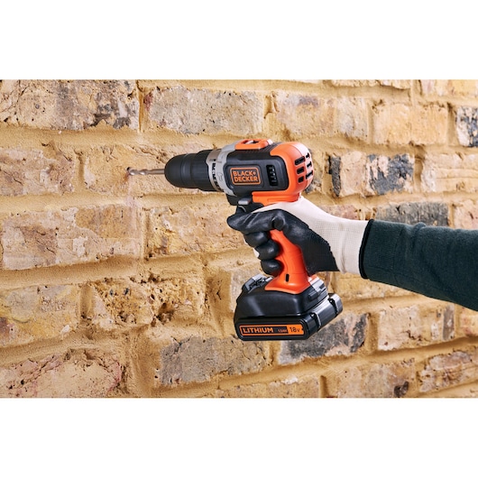Perceuse sans fil BLACK+DECKER BCD003C2K utilisée sur un mur en briques, design orange et noir, main gantée tenant l'outil.
