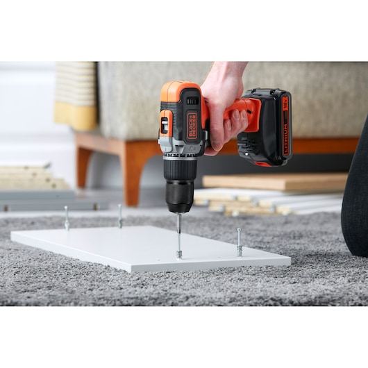 BLACK+DECKER BCD003ME2K perceuse sans fil en usage, vissant des vis dans une planche blanche sur moquette.