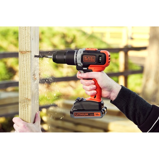 Perceuse sans fil BLACK+DECKER BCD003ME2K 18V utilisée pour percer du bois en extérieur, étincelles et deux mains visibles.