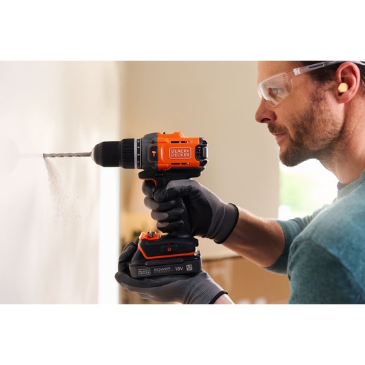 BLACKDECKER BCD383D1 snoerloze boormachine met zwart-oranje behuizing, gehandschoende hand boort in een muur.