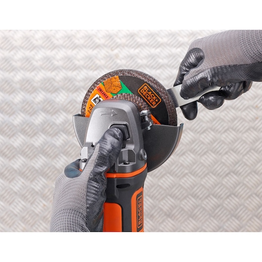 Foto van BLACKDECKER BCG720N haakse slijper die wordt afgesteld met gehandschoende handen, close-up van gereedschap en schijf.