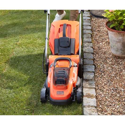 Tondeuse à gazon orange sans fil BLACKDECKER BCMW33185L2 tondant l’herbe près d’une bordure de gravier, vue de dessus.