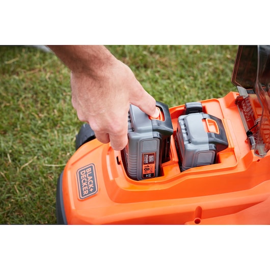 Photo du tondeuse à gazon BLACKDECKER BCMW33185L2 avec main insérant la batterie, orange et gris, fond d’herbe extérieur.