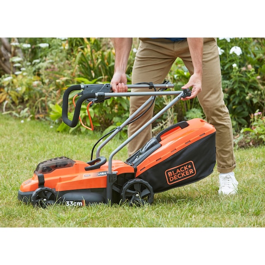 Photo de la tondeuse sans fil BLACK+DECKER BCMW33185L2 utilisée sur une pelouse, orange et noire avec bac de ramassage.