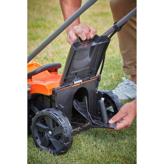 Photo de la tondeuse BLACK+DECKER BCMW33185L2, gros plan sur l’arrière avec une personne ouvrant le capot de décharge sur l’herbe.