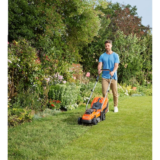 Photo d'une personne utilisant la tondeuse orange BLACK+DECKER BCMW3318N sur l'herbe dans un jardin verdoyant.