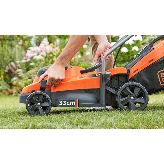 Photo de la tondeuse BLACK+DECKER BCMW3318N, orange et noire, vue latérale, utilisateur ajustant la poignée, largeur de coupe 33 cm.