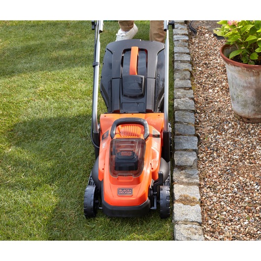 BLACKDECKER BCMW3336N tondeuse vue de face sur pelouse à côté d’une bordure en pierre et pot de fleurs.