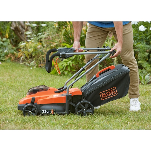 Photo de la tondeuse BLACK+DECKER BCMW3336N, design noir et orange, poignée pliable et bac de ramassage d’herbe utilisée sur la pelouse.