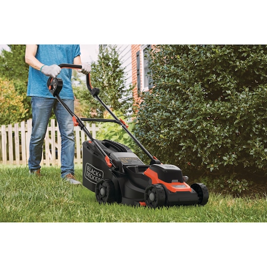 Een persoon maait het gras met een draadloze grasmaaier van BLACK+DECKER in een tuin bij een woning. De maaier is zwart en oranje, en de persoon draagt handschoenen en een spijkerbroek.