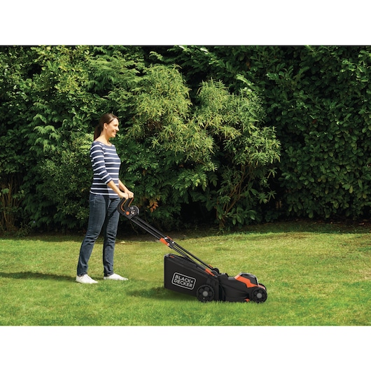 Een persoon gebruikt een BLACK+DECKER grasmaaier om het gras in een tuin te maaien, met dichte groene struiken op de achtergrond.