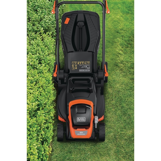 Bovenaanzicht van een BLACK+DECKER 18V draadloze grasmaaier op een gazon, met het handvat, het bedieningspaneel en een deel van een gesnoeide haag aan de linkerkant.