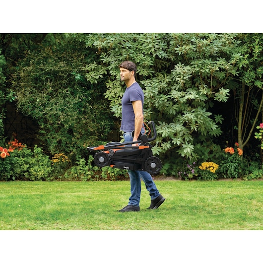 Een persoon met een vervaagd gezicht draagt een opgevouwen BLACK+DECKER grasmaaier door een grasrijke tuin met weelderig groen en bloemen op de achtergrond.