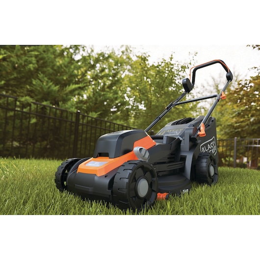 Een BLACK+DECKER grasmaaier met een oranje en zwart ontwerp staat op groen gras in een tuin. Bomen en een metalen hek zijn zichtbaar op de achtergrond.