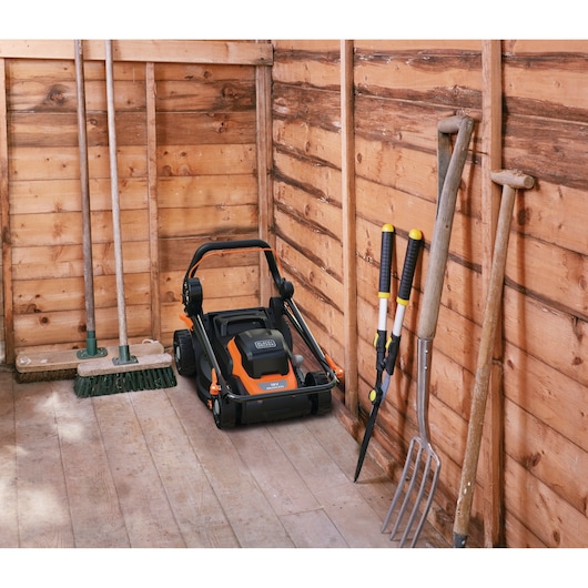 Een BLACK+DECKER grasmaaier opgeslagen in een houten tuinhuisje, omringd door verschillende tuingereedschappen zoals twee bezems, een hooivork, een heggenschaar en twee schoppen die tegen de houten wanden staan.