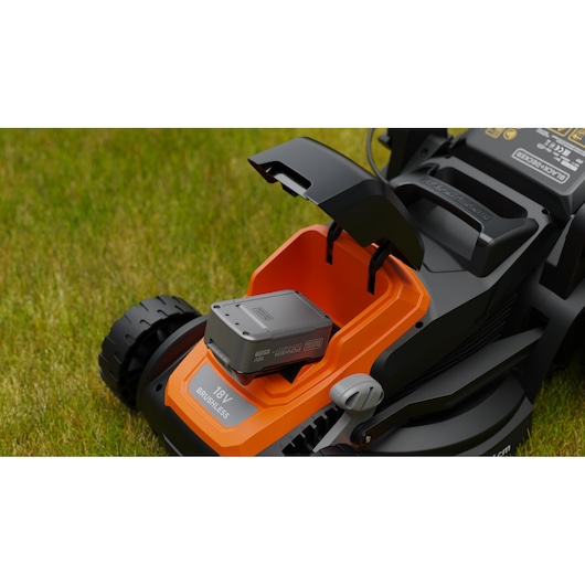 Close-up van een draadloze BLACK+DECKER grasmaaier op gras, met het batterijcompartiment open en een 18V batterij zichtbaar binnenin.
