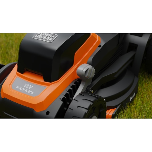Close-up van een BLACK+DECKER 18V borstelloze grasmaaier, met een oranje en zwarte behuizing, verstelbare hoogtehendel en wiel, geplaatst op groen gras.
