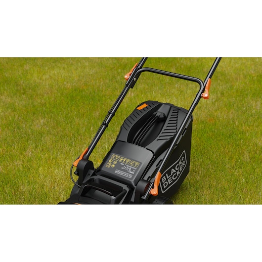 Close-up van een BLACK+DECKER grasmaaier op groen gras, te zien zijn de hendel, het bedieningspaneel met waarschuwingslabels en de gemerkte grasopvangbak.