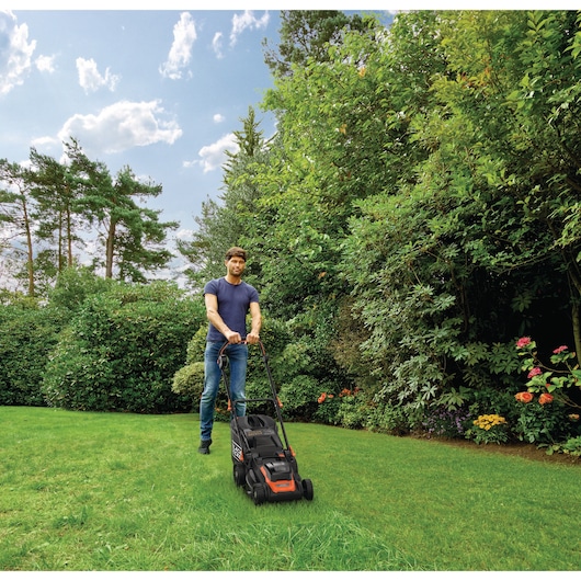 Een persoon met een vervaagd gezicht maait het gazon met een BLACK+DECKER grasmaaier in een tuin omringd door groene struiken en bomen onder een blauwe lucht.