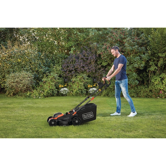 Een persoon gebruikt een BLACK+DECKER grasmaaier op een gazon in een tuin, omringd door struiken en bloemen.