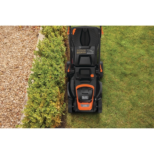 Bovenaanzicht van een BLACK+DECKER grasmaaier die gras maait naast een haag en een grindpad.