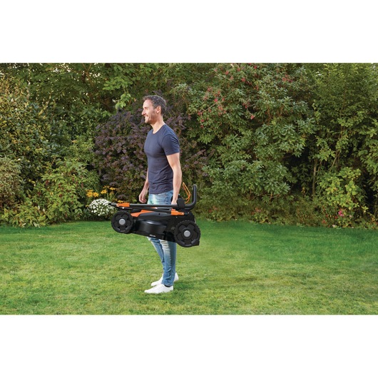 Een persoon draagt een opgevouwen BLACK+DECKER grasmaaier over een groene grasveld in een tuin, omringd door struiken en planten. Het gezicht van de persoon is wazig gemaakt.