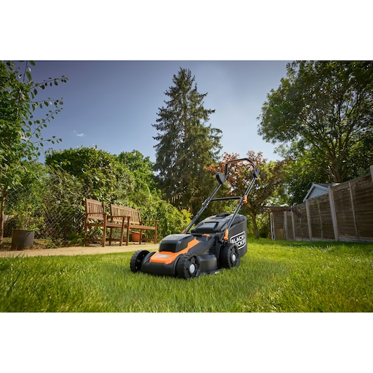 Een BLACK+DECKER grasmaaier staat op een groen gazon in een achtertuin. De scène bevat bomen, struiken, een houten bank en een houten hek onder een heldere blauwe lucht.