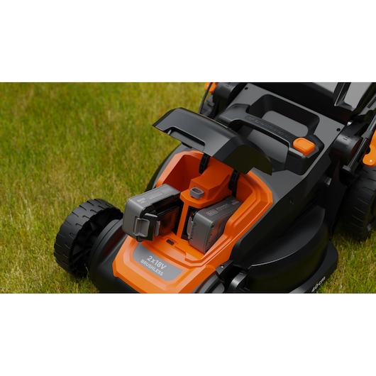 Close-up van een BLACK+DECKER grasmaaier met twee oplaadbare accu’s die in het accuvak zijn geplaatst. De maaier staat op groen gras en het oranje-zwarte ontwerp is zichtbaar.