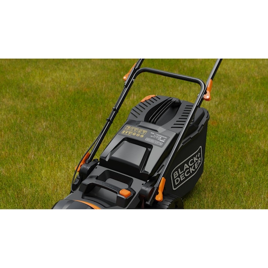 Een close-up van een BLACK+DECKER grasmaaier op groen gras. De maaier is zwart met oranje accenten en heeft een zichtbare hendel en een opvangbak voor gras.