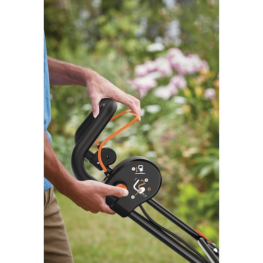 Close-up van een persoon die de bedieningshendel van een BLACK+DECKER tuingereedschap afstelt, met een tuin op de achtergrond met groen blad en paarse bloemen.