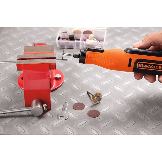 BLACK+DECKER BCRT8IK roterend gereedschap in gebruik, vastgehouden bij rode bankschroef met accessoires op metalen oppervlak.