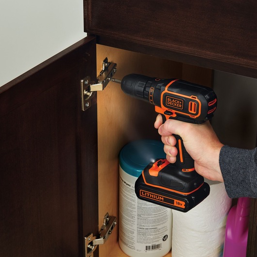 BLACK+DECKER BDCDC18K1B perceuse sans fil utilisée pour installer une charnière d'armoire, design noir et orange, vue rapprochée.