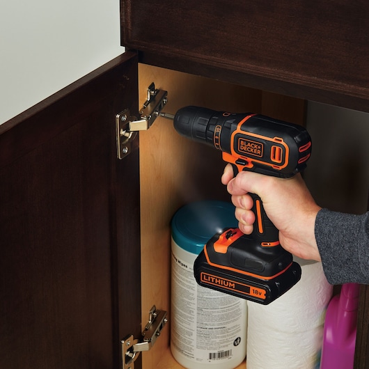 BLACKDECKER BDCDC18KB perceuse sans fil, orange et noire, utilisée pour fixer une charnière d’armoire en intérieur.