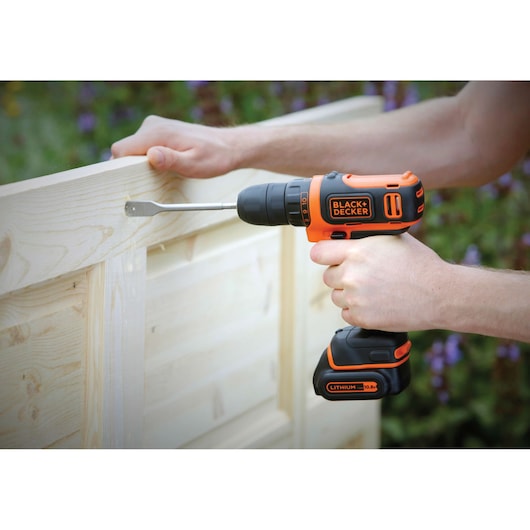 Perceuse sans fil BLACK+DECKER BDCDD12 utilisée sur du bois clair, couleurs orange et noir, tenue par une personne.