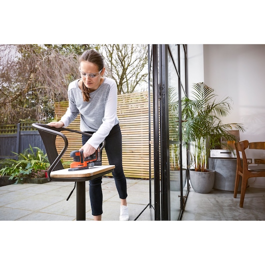 Photo de la ponceuse BLACKDECKER BDCDS18 utilisée pour poncer une chaise en extérieur près d’une porte-fenêtre.