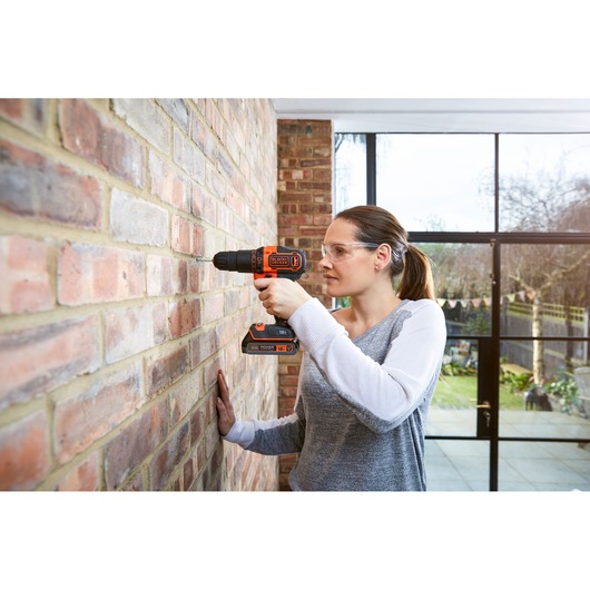 BLACK+DECKER BDCHD18S2KA perceuse sans fil utilisée sur mur en briques, détails orange et noirs visibles.