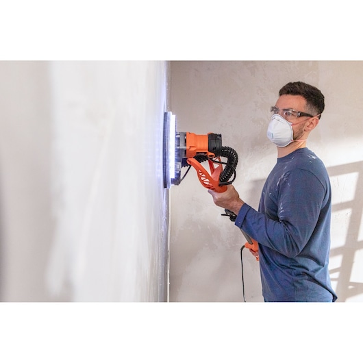 Persoon gebruikt BLACK+DECKER wandschuurmachine op witte muur, draagt masker en bril voor veiligheid.
