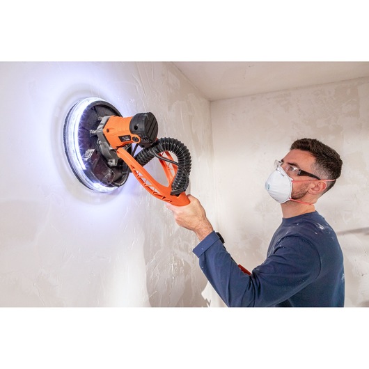 Persoon gebruikt BLACK+DECKER elektrische muur schuurmachine met LED-licht, draagt masker en bril.