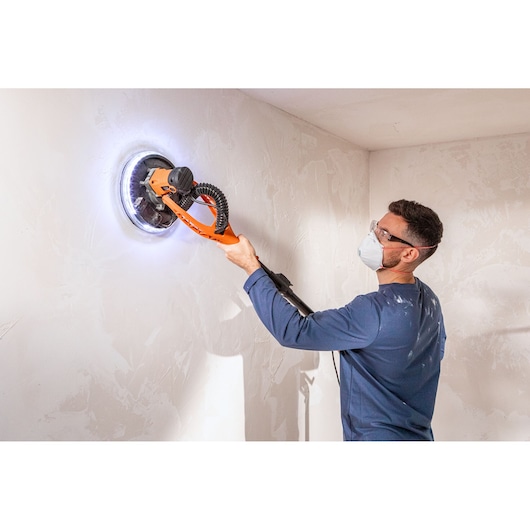 Persoon gebruikt een BLACK+DECKER muurschuurmachine met LED-licht, draagt een masker en bril.