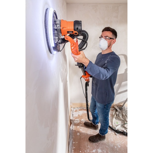 Persoon schuurt muur met BLACK+DECKER wand-schuurmachine met LED-licht en draagt masker en bril.