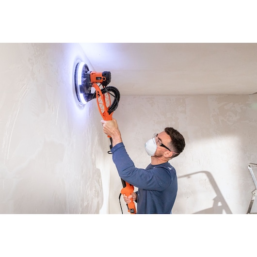 Persoon schuurt plafond met BLACK+DECKER LED gipsplaten schuurmachine, draagt masker en oogbescherming.