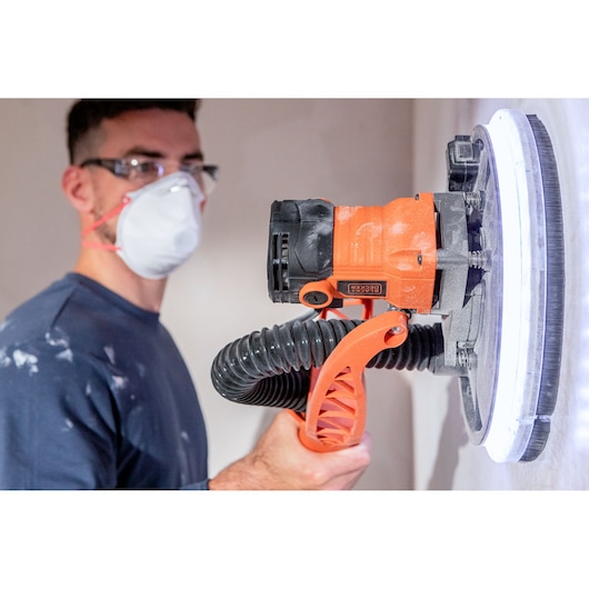 Persoon gebruikt BLACK+DECKER wandschuurmachine met masker en bril voor renovatie binnenshuis.