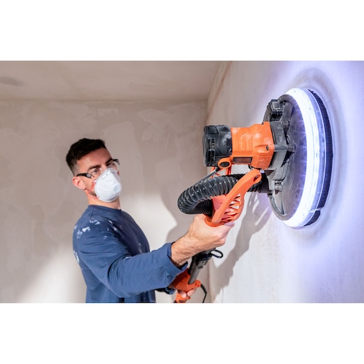 Persoon gebruikt BLACK+DECKER wandenschuurmachine met LED-licht, draagt masker voor bescherming tegen stof.