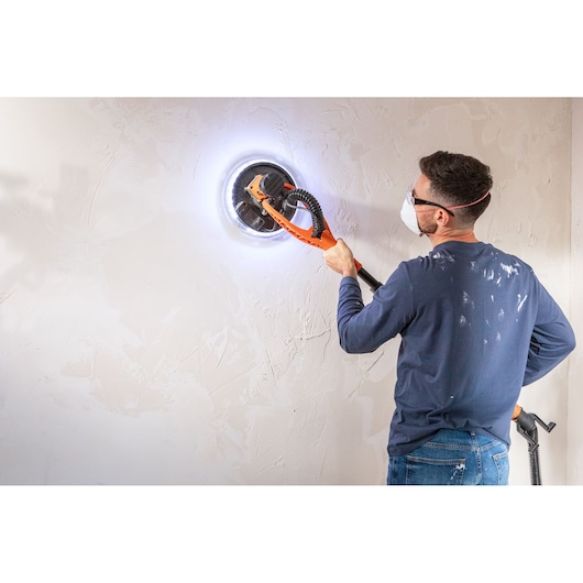 Persoon schuurt witte muur met BLACK+DECKER schuurmachine met LED, draagt veiligheidsbril en masker.