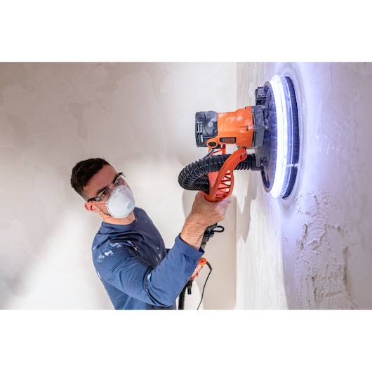 Persoon gebruikt BLACK+DECKER muurschuurmachine met LED-licht op gips, draagt masker en bril.