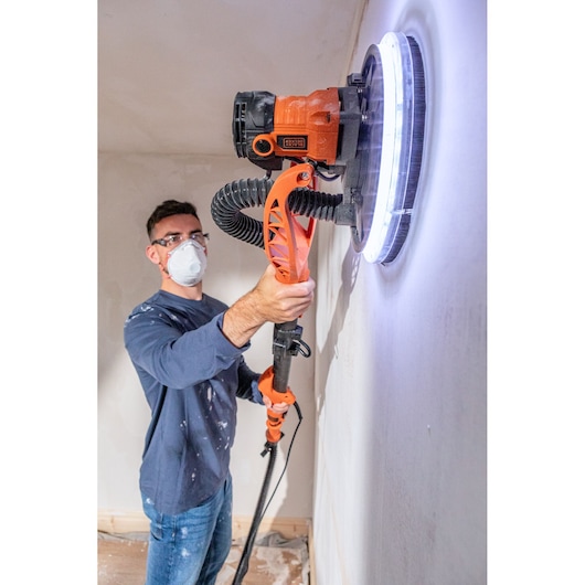 Persoon schuurt muur met BLACK+DECKER elektrische schuurmachine, draagt masker tegen stof bij renovatie.