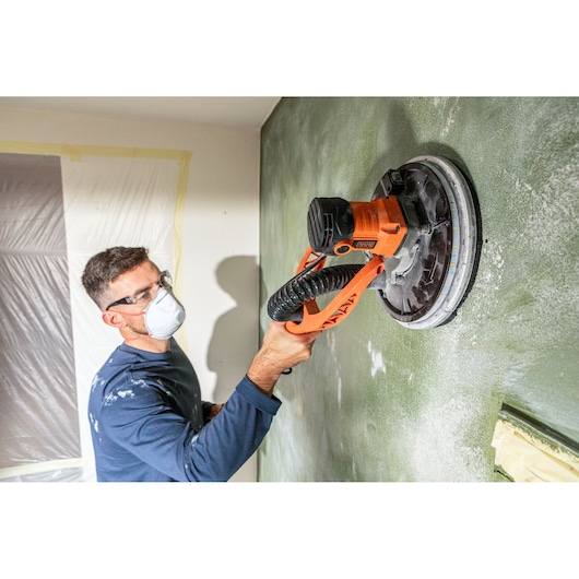 Persoon gebruikt een BLACK+DECKER elektrische wandenschuurmachine met slang op een groene muur bij renovatie.